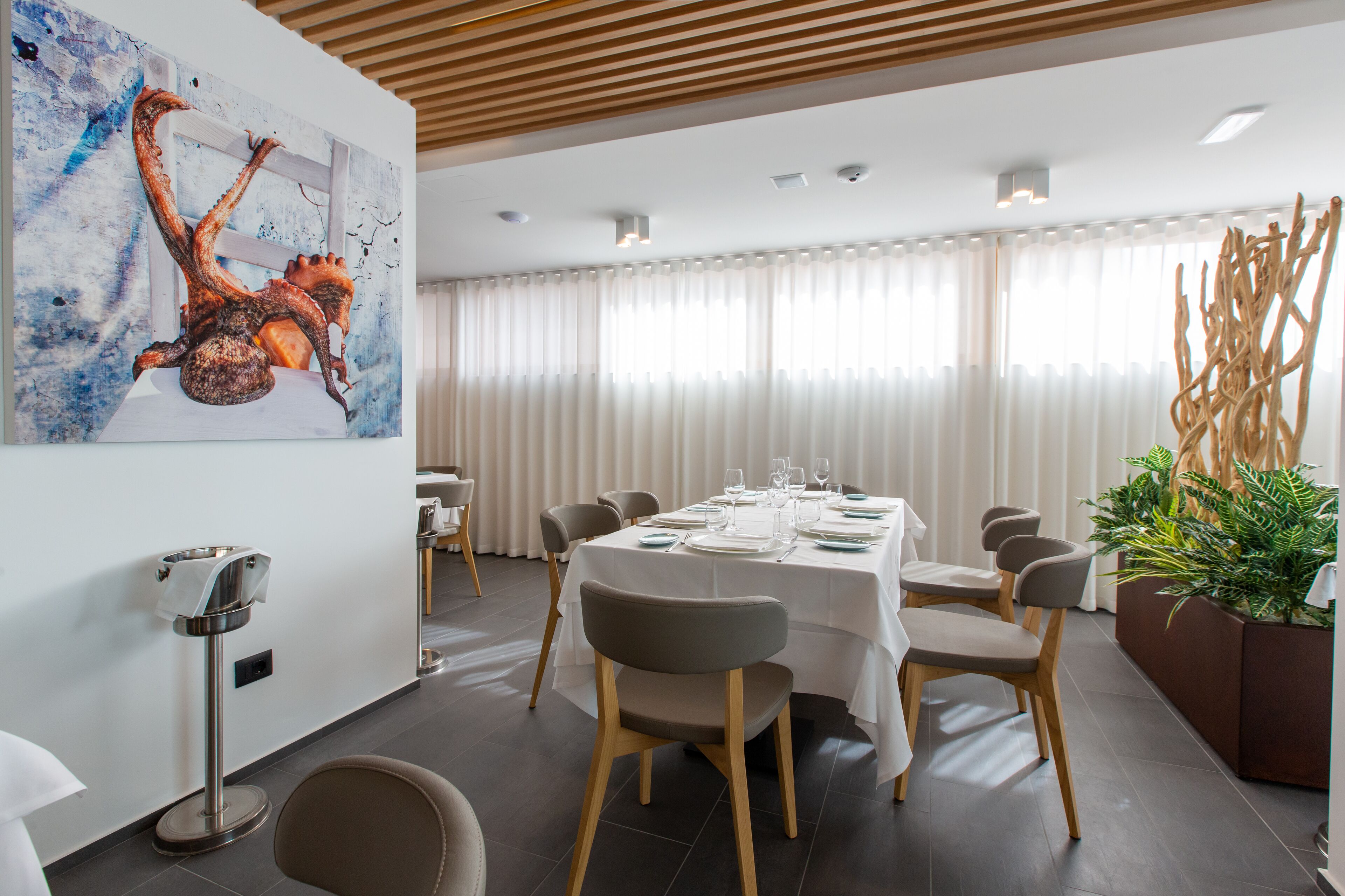 Un espace repas moderne avec une table blanche dressée pour six personnes, entourée de chaises aux pieds en bois. Un grand tableau abstrait orne le mur, ainsi qu’un lampadaire et une plante en pot placés sur le côté.