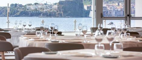 A restaurant with tables set for dining, overlooking a coastal town with a historic castle.