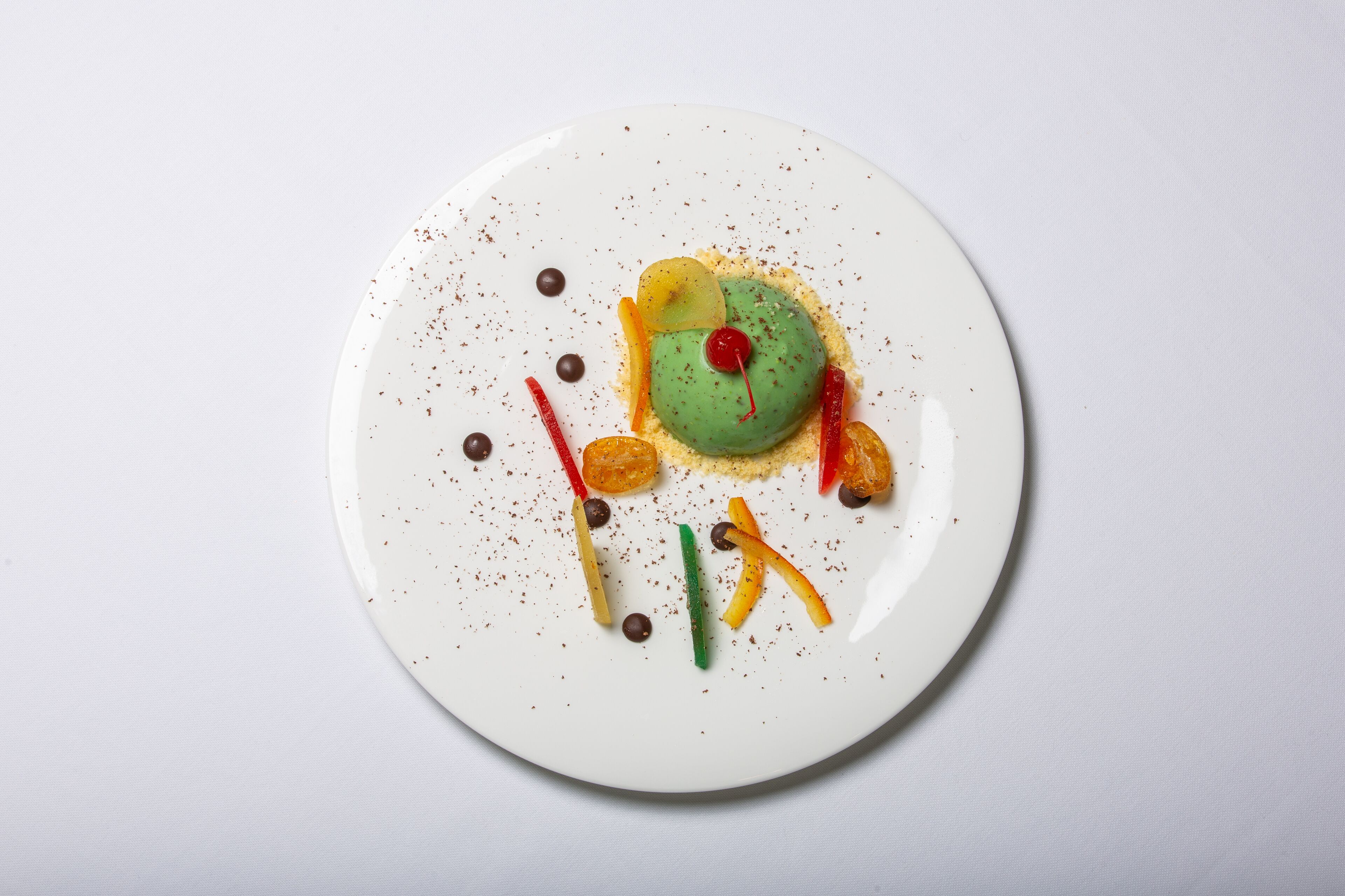 Ein weißer Teller mit einem grünen Dessert, umgeben von bunten Garnierungen und Schokoladenstückchen.