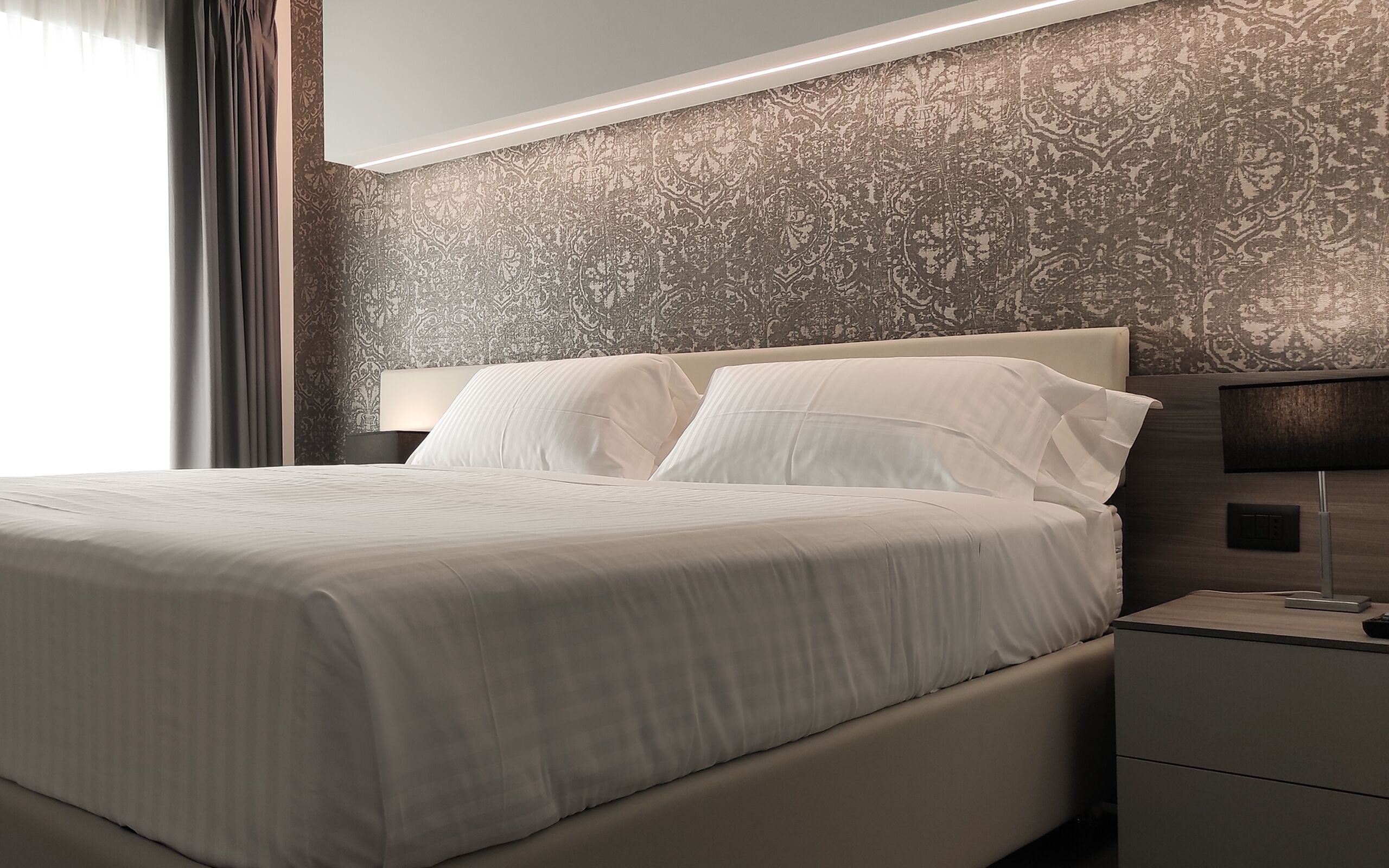 Quarto de hotel moderno com uma cama grande, uma mesa de cabeceira com abajur e papel de parede estampado.