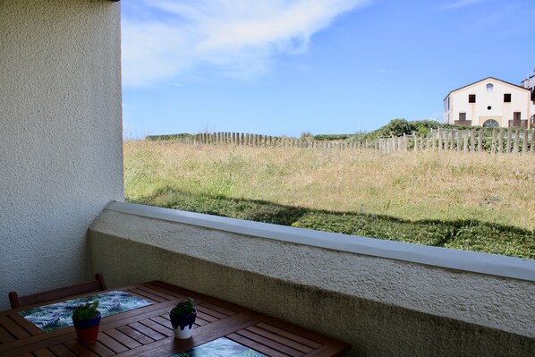 Un balcon avec une table et des chaises en bois, un pot de fleurs et une vue sur un champ verdoyant et un bâtiment situé au loin.