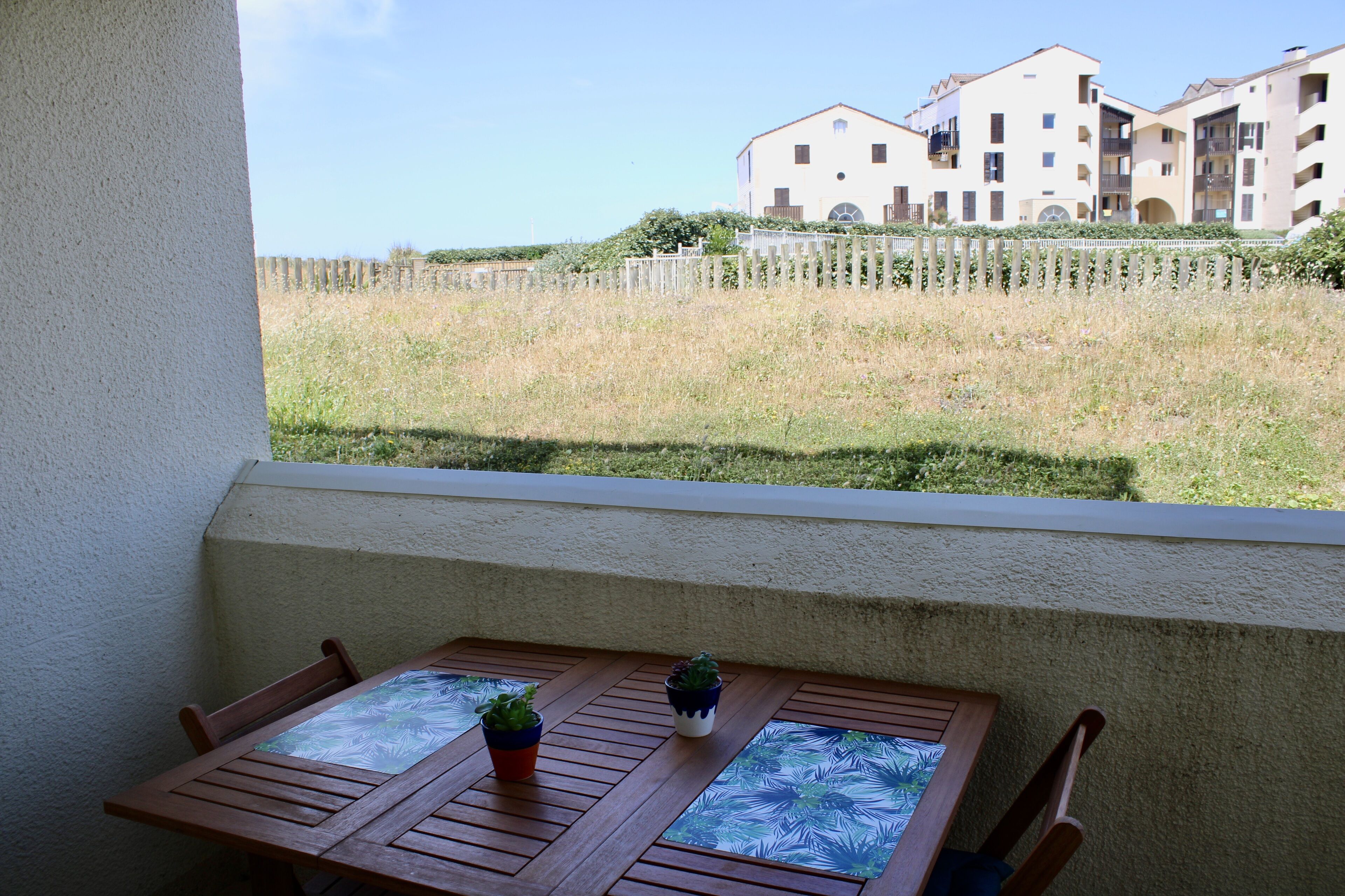 Une table en bois, ornée de deux plantes en pot, se trouve sur un balcon qui donne sur un espace verdoyant et une rangée de bâtiments.