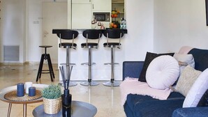 A modern living room with a dark blue sofa, three black bar stools, a small round table with candles and a plant, and a kitchen area with a microwave and fruit basket in the background. - Tranquil King David Apartment with Free Parking (Jerusalem)