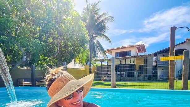 A person relaxing by a pool with a clear view of a building and palm trees in the background. - Pousada Trilha do Mar (Marechal Deodoro)