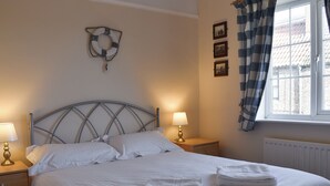 Un lit bien fait, avec du linge de lit blanc, deux lampes de chevet, une horloge murale et une fenêtre ornée de rideaux à carreaux.