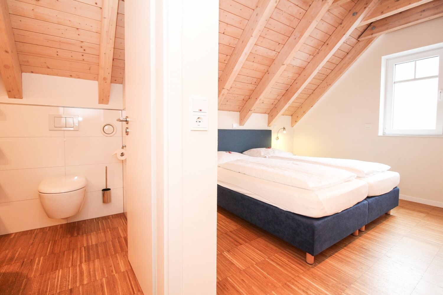 Un dormitorio con techo de madera, una cama con ropa de cama blanca y un ventanal que deja entrar luz natural.