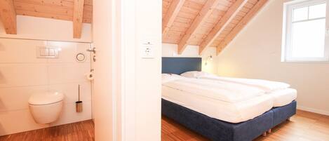 Un dormitorio con techo de madera, una cama con ropa de cama blanca y un ventanal que deja entrar luz natural.