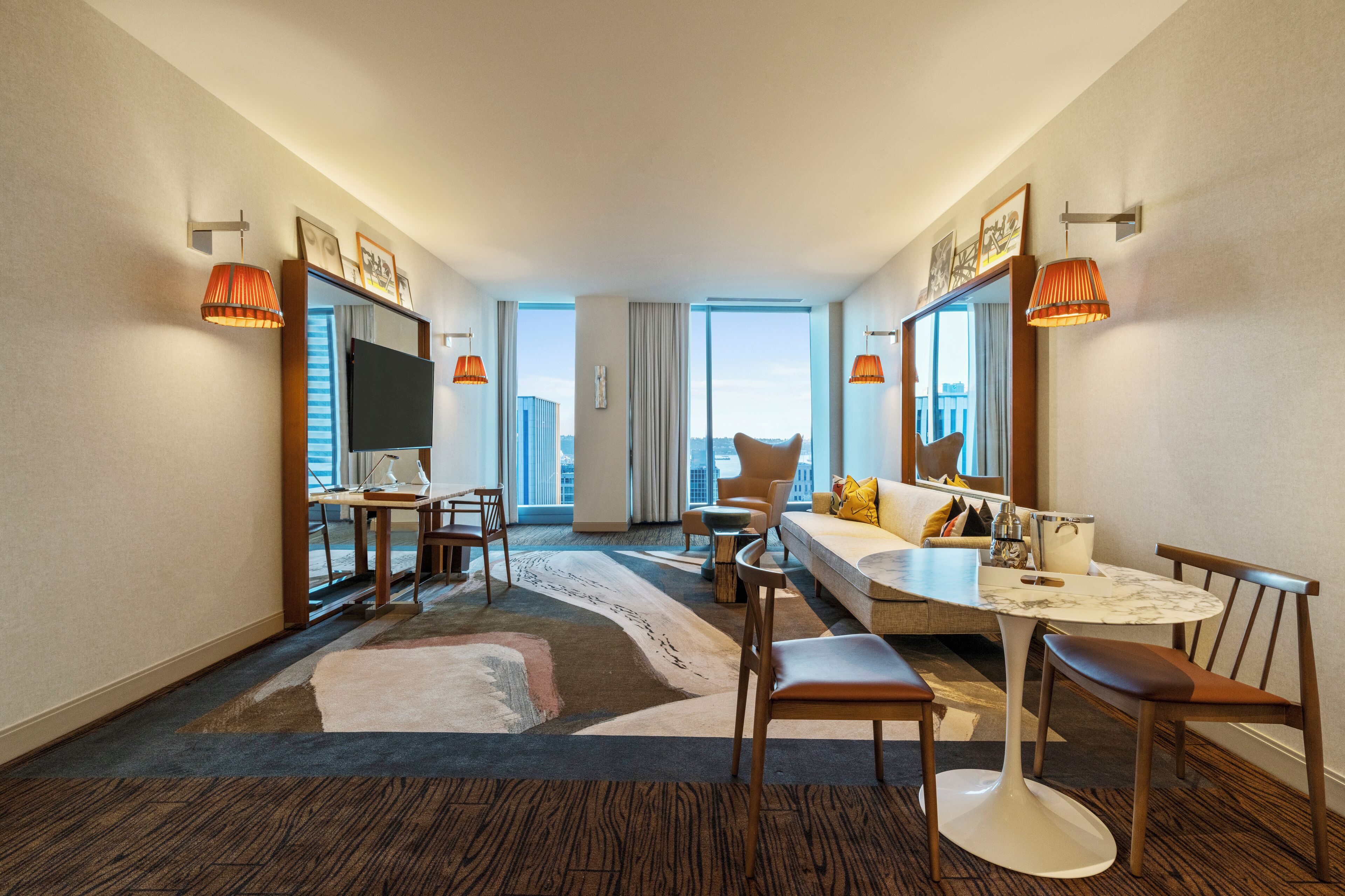 A hotel room with a sofa, a dining table, a TV, and a large window offering a city view.