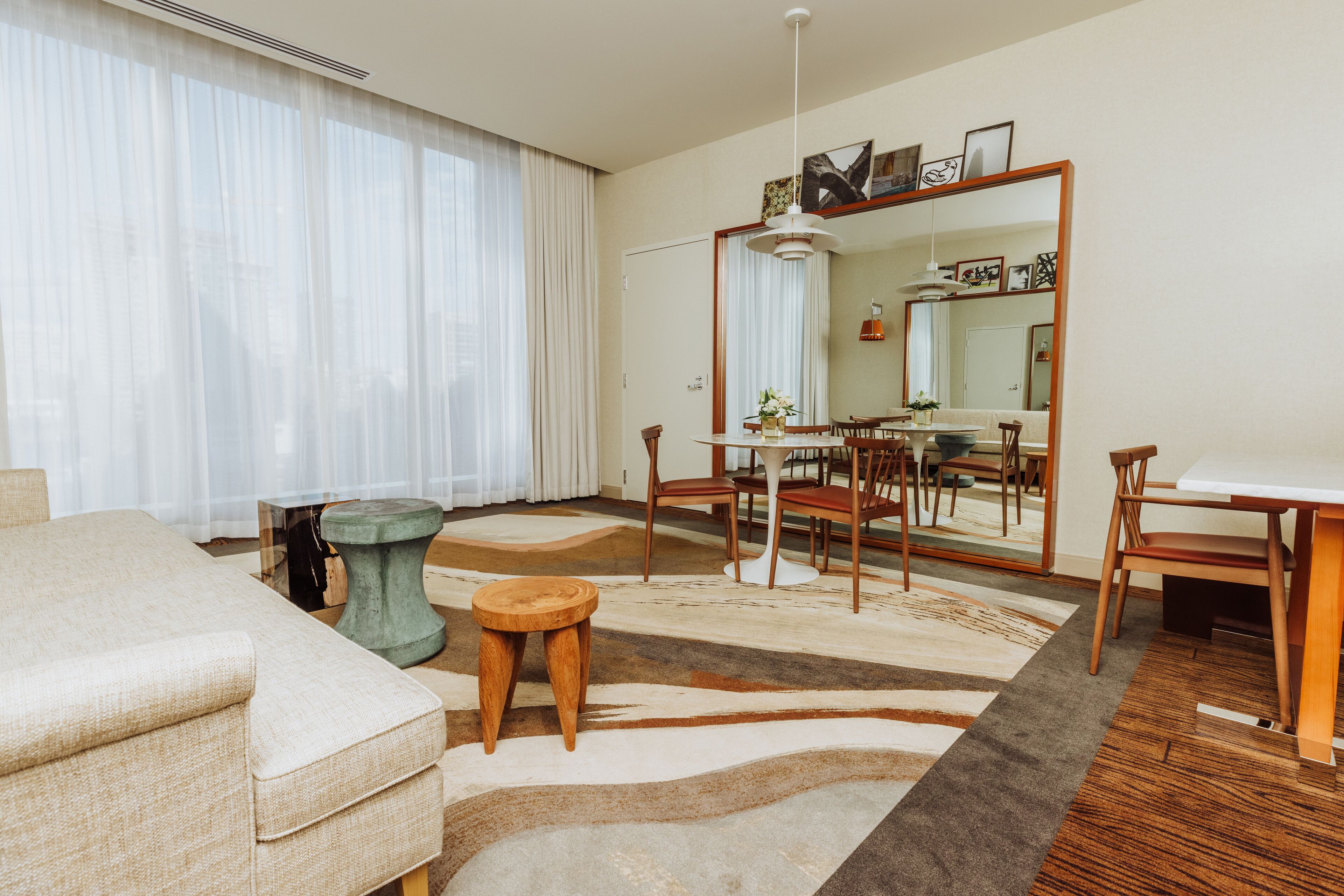 Una habitación de hotel moderna con una cama grande, una mesa de comedor, sillas, un taburete pequeño y un espejo amplio.