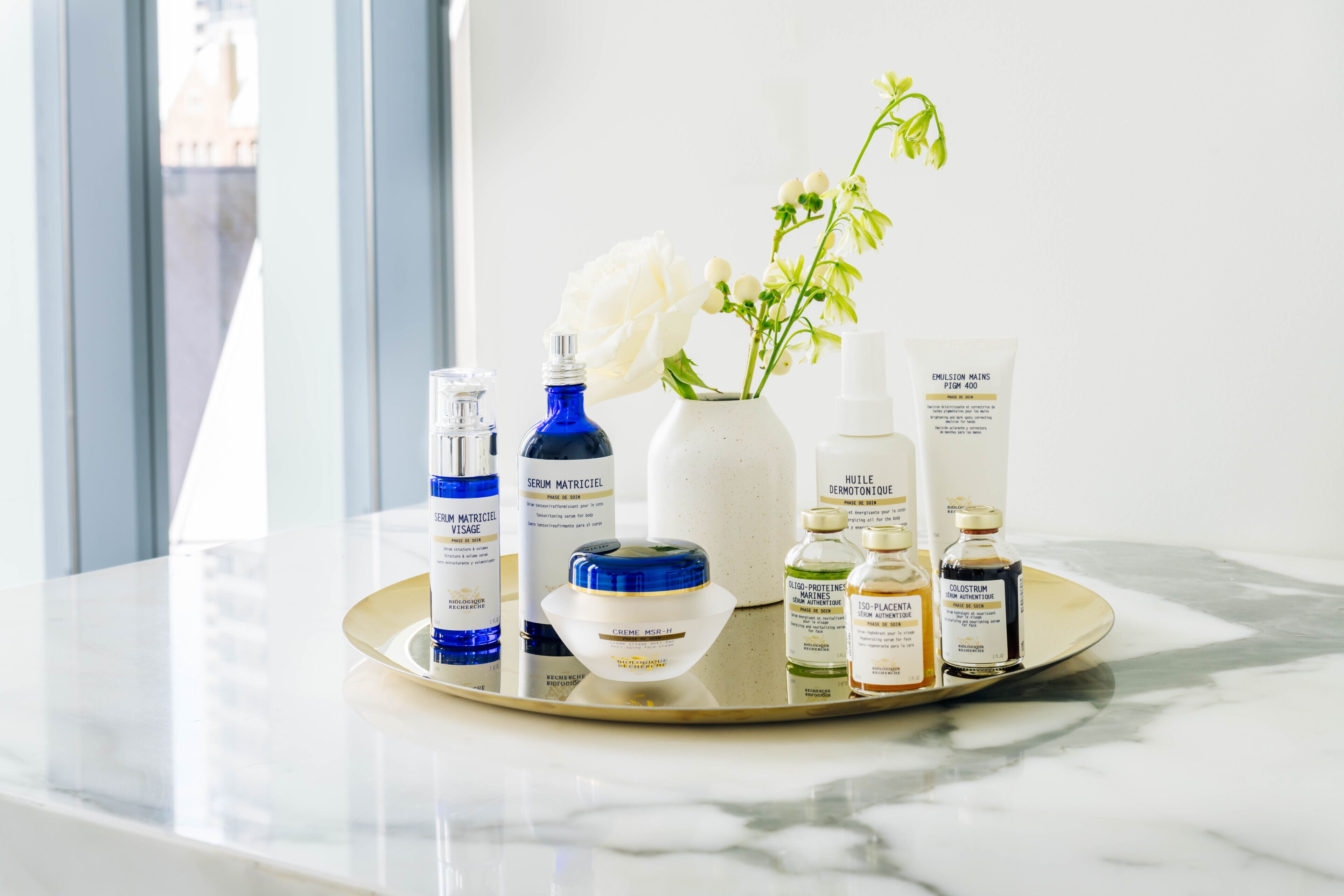 A tray with various skincare products on a marble countertop near a window.