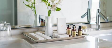 Un tocador de baño con encimera de mármol blanco, un espejo, un jarrón blanco con flores blancas y una bandeja con tres botellas pequeñas.