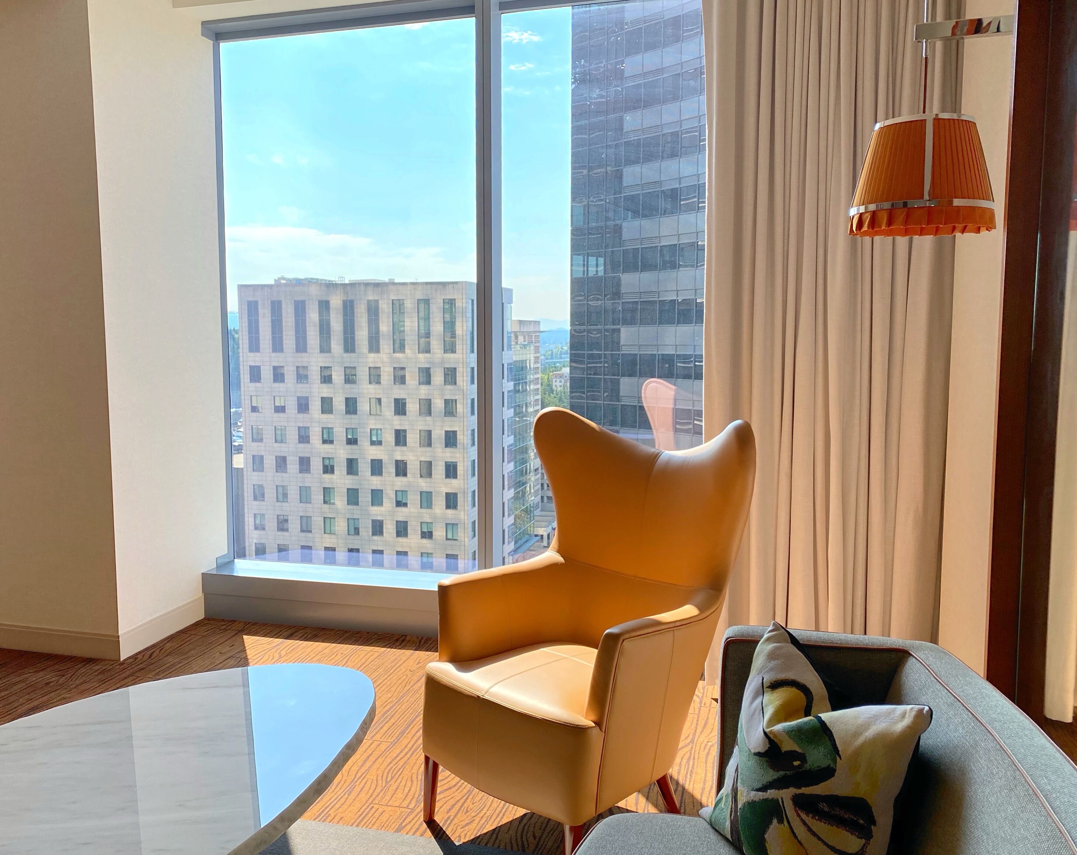 Une chambre d’hôtel moderne avec une grande fenêtre, un fauteuil jaune, un canapé gris et une petite table.
