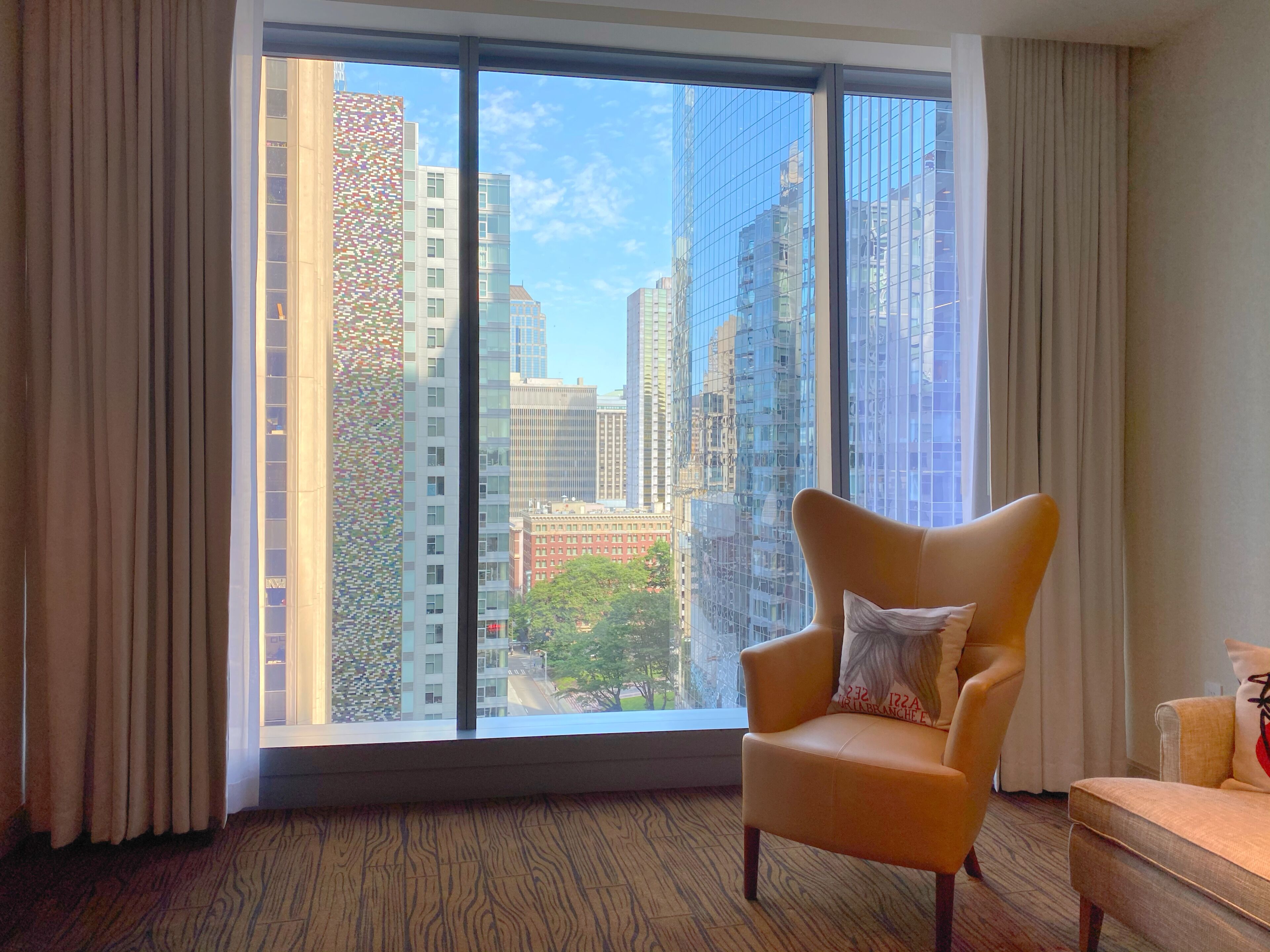 A modern hotel room with a large window offering a city view, a comfortable armchair, and a small sofa.