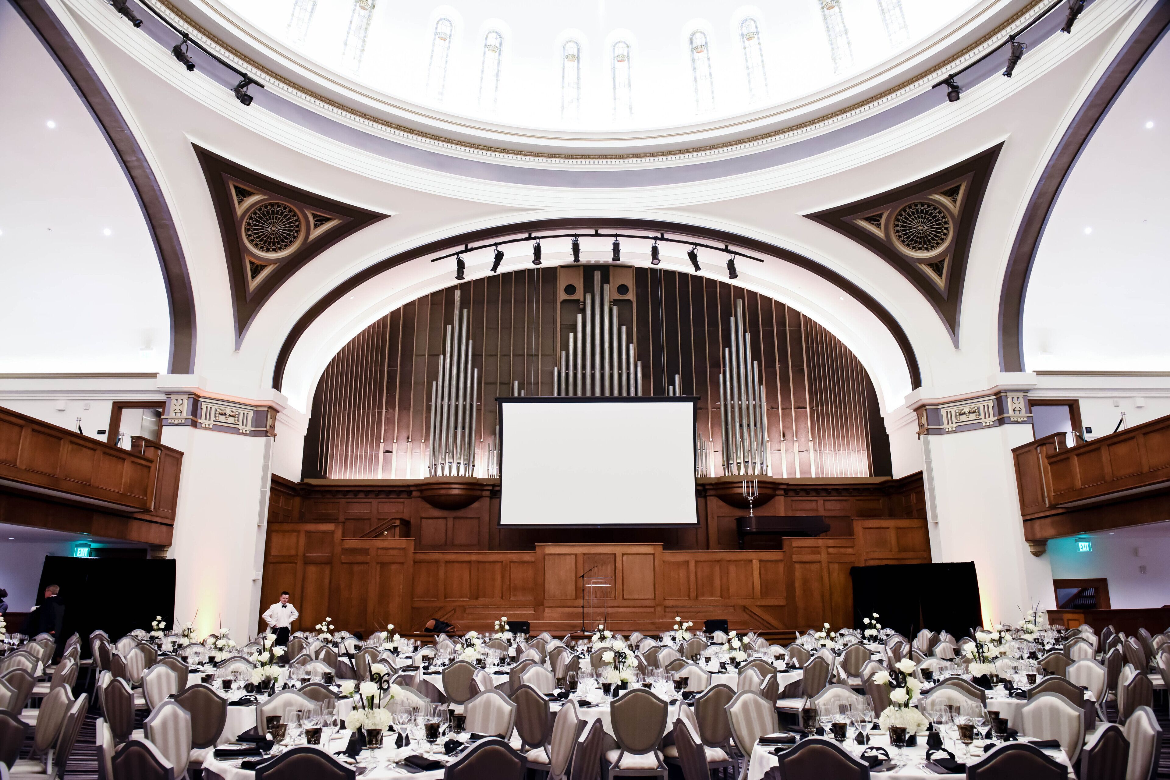 A large conference room with tiered seating, a stage with a large screen, and a decorative organ.