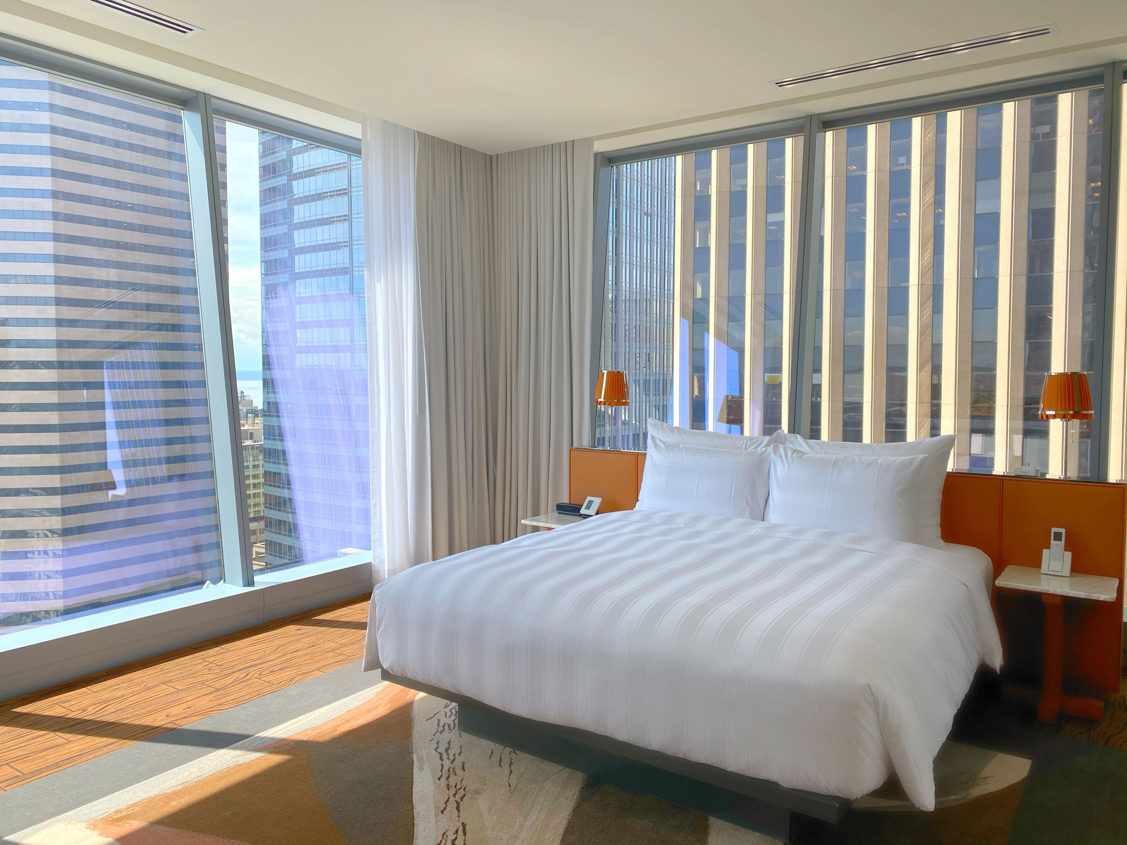 Une chambre d’hôtel avec un grand lit, une table de chevet, deux lampes et une vue sur le paysage urbain grâce à de grandes fenêtres du sol au plafond.