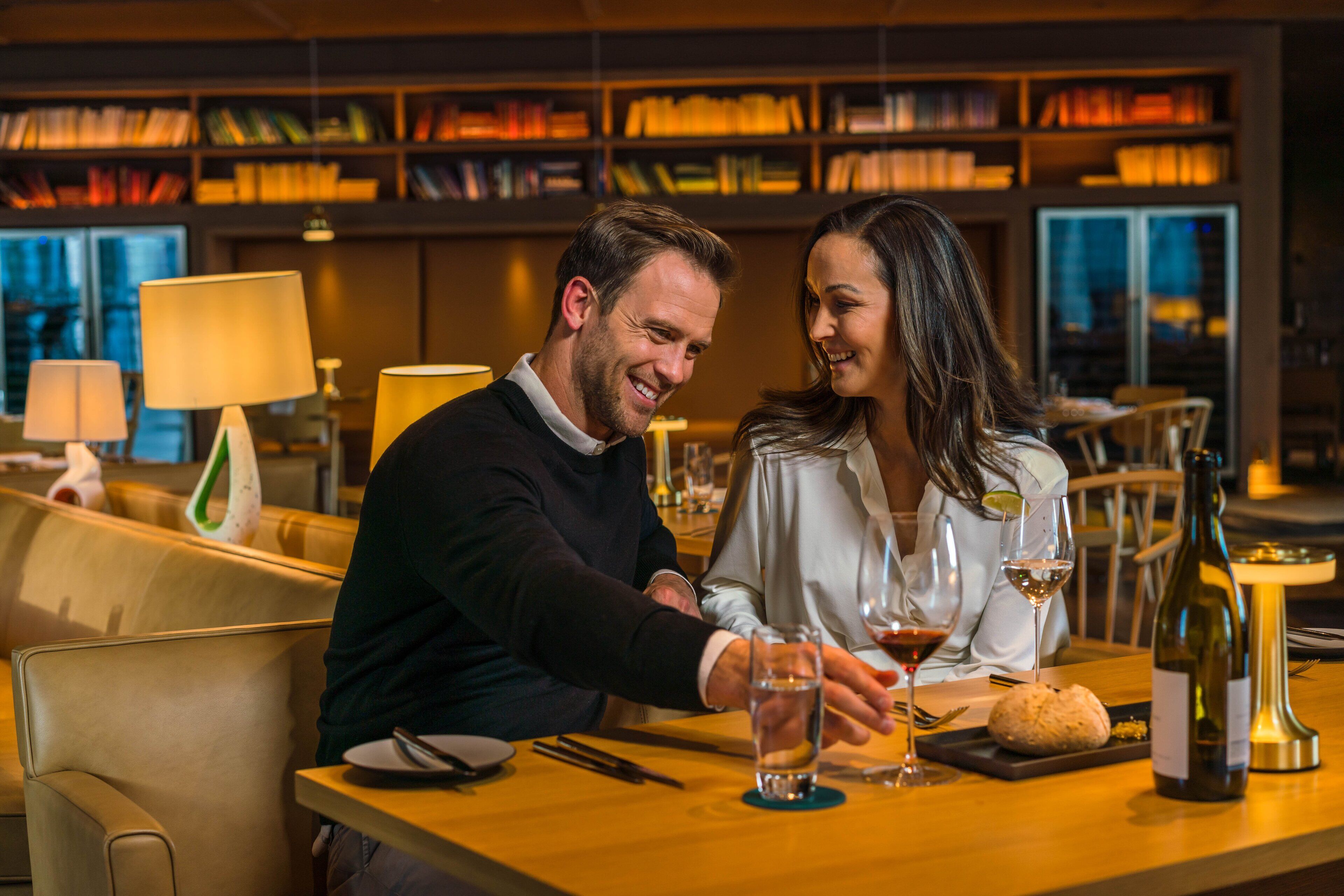 A couple dining at a table with a bottle of wine, a glass of water, and a loaf of bread.