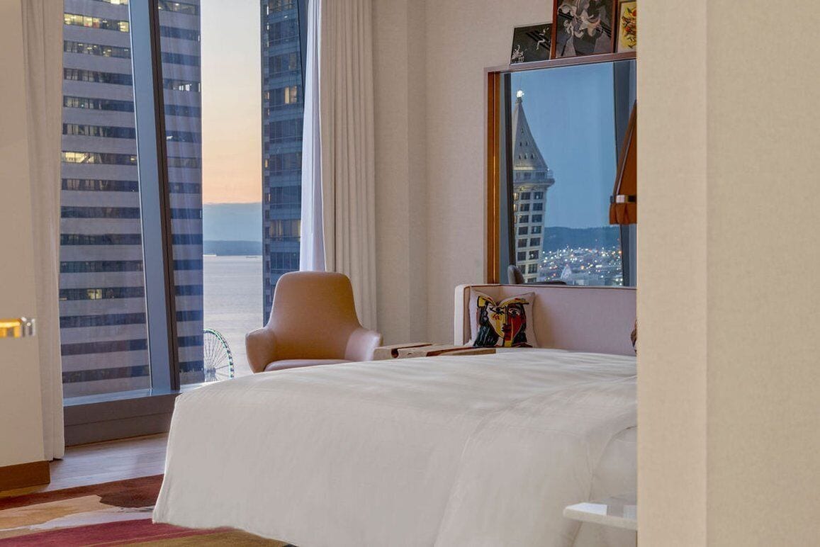 A hotel room with a large bed, a chair, a desk, and a view of the cityscape through the window.