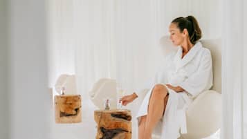 A person seated in a white chair in a spa-like setting with a wooden stool and a glass of water.