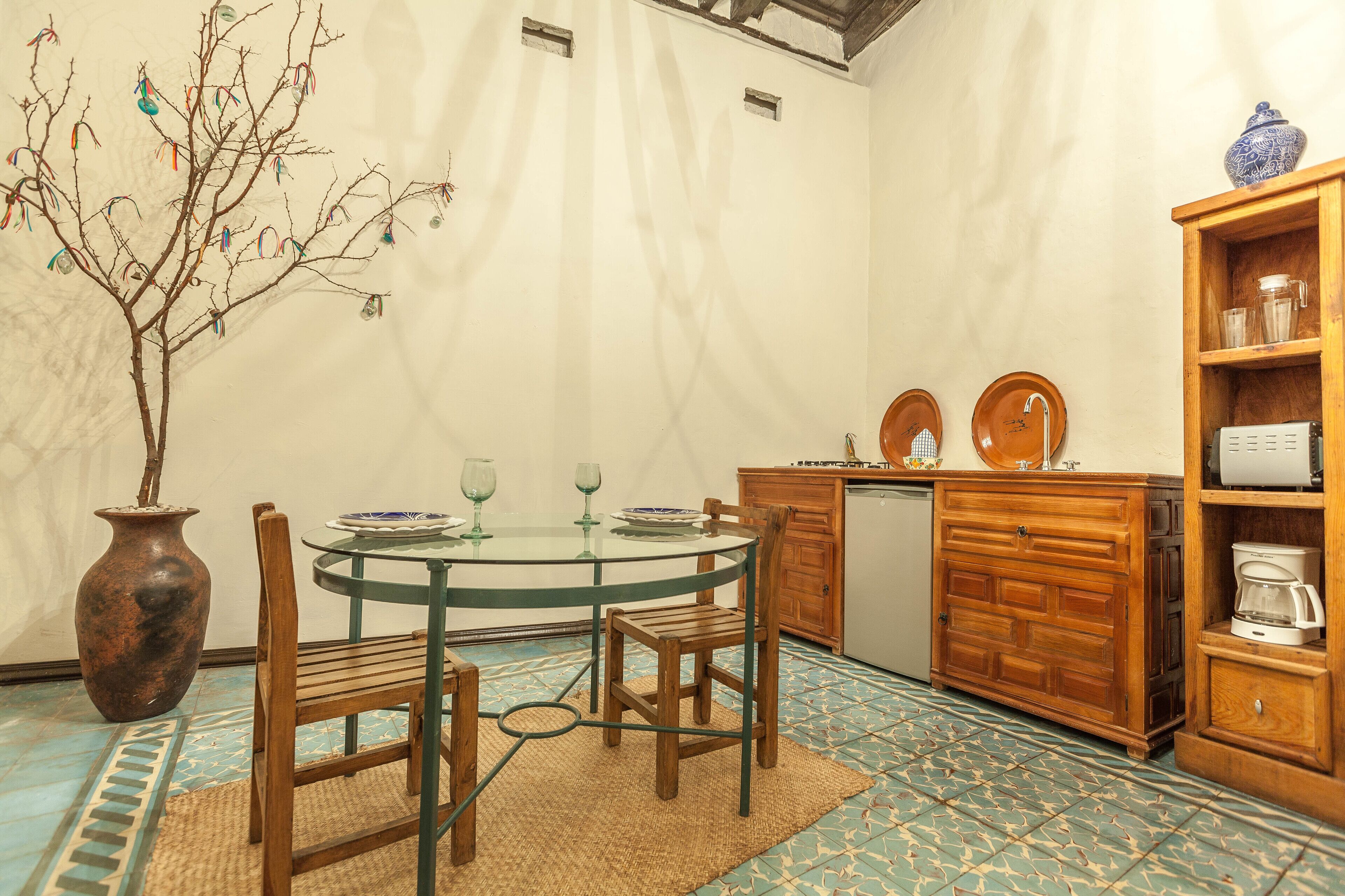 A dining area with a round glass table, wooden chairs, a vase with branches, and a cabinet with kitchen items.
