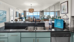 A modern kitchen with a black countertop, stainless steel sink, and teal cabinets. There is a dining area with a view of the ocean. - Beach Front, Renovated Kitchen, Free Beach Chair Service (Miramar Beach)