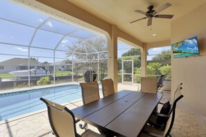 A poolside dining area with a table and chairs, a ceiling fan, and a view of a swimming pool and a boat.