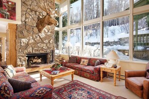 Un salon chaleureux avec une cheminée en pierre, de grandes fenêtres donnant sur un paysage enneigé et des meubles aux motifs originaux.
