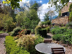 Un jardín con una mesa metálica redonda y bancos, rodeado de diversas plantas y un edificio de piedra.