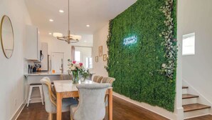 Een eetruimte met een houten tafel en stoelen, een groene muur met een neonbord, en een trap.