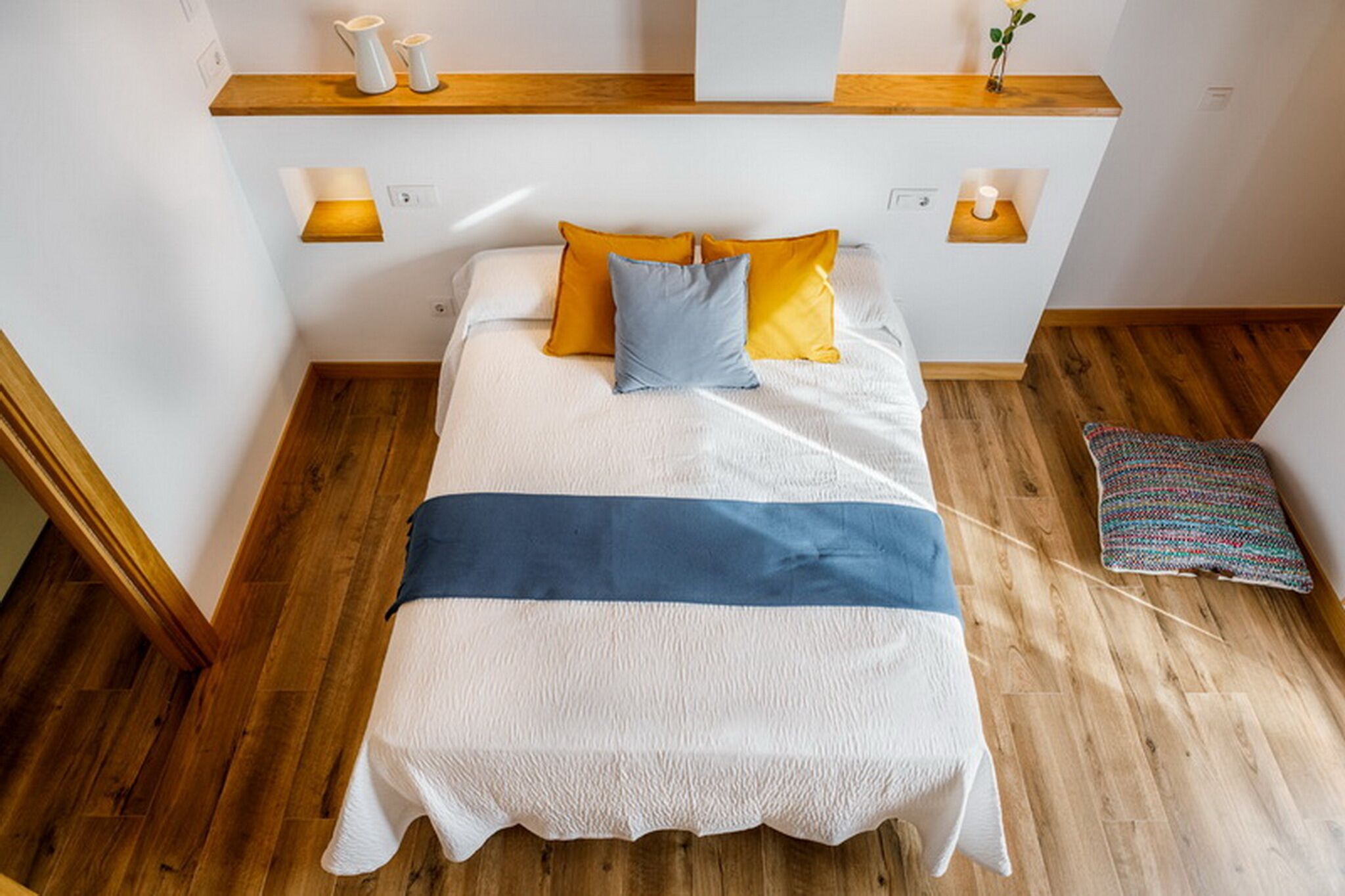 Una cama bien tendida con una colcha blanca y azul, dos almohadas en amarillo y azul, y un suelo de madera.