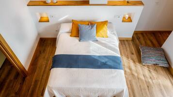 Una cama bien tendida con una colcha blanca y azul, dos almohadas en amarillo y azul, y un suelo de madera.