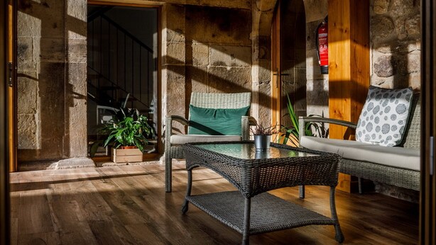 A cozy room with a wicker sofa, a small table, and potted plants. A stone wall and a wooden door are visible. - Posada Rural Fuente Juliana (Ruente)