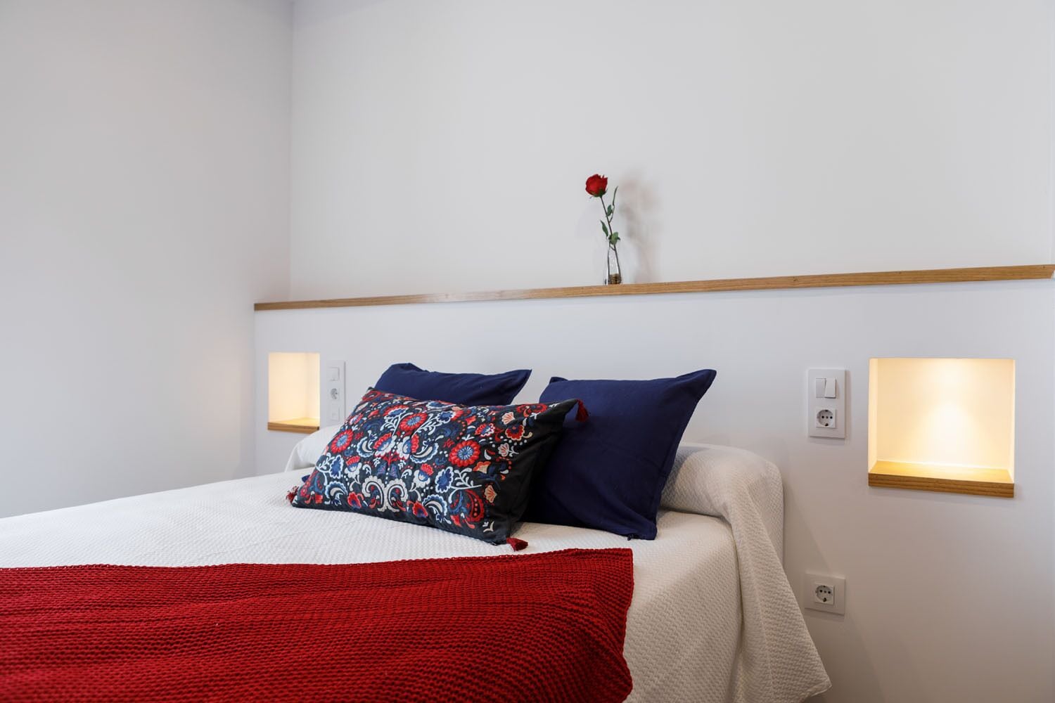 Una cama bien hecha con una colcha roja, dos almohadas y una almohada decorativa con estampado floral. Sobre la cama, en un estante, hay una única rosa roja.