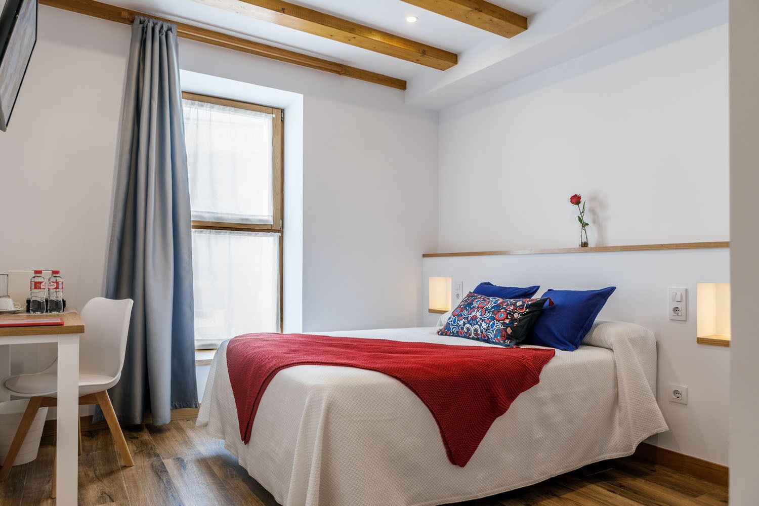 A bedroom with a bed, a desk with a chair, a window with curtains, and a bedside table with a vase of flowers.