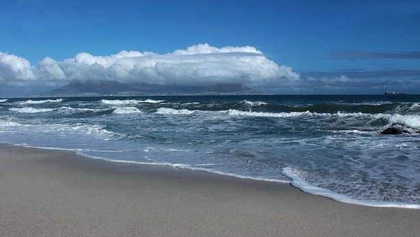 Une plage où les vagues se brisent sur le rivage, un ciel dégagé et une montagne lointaine.