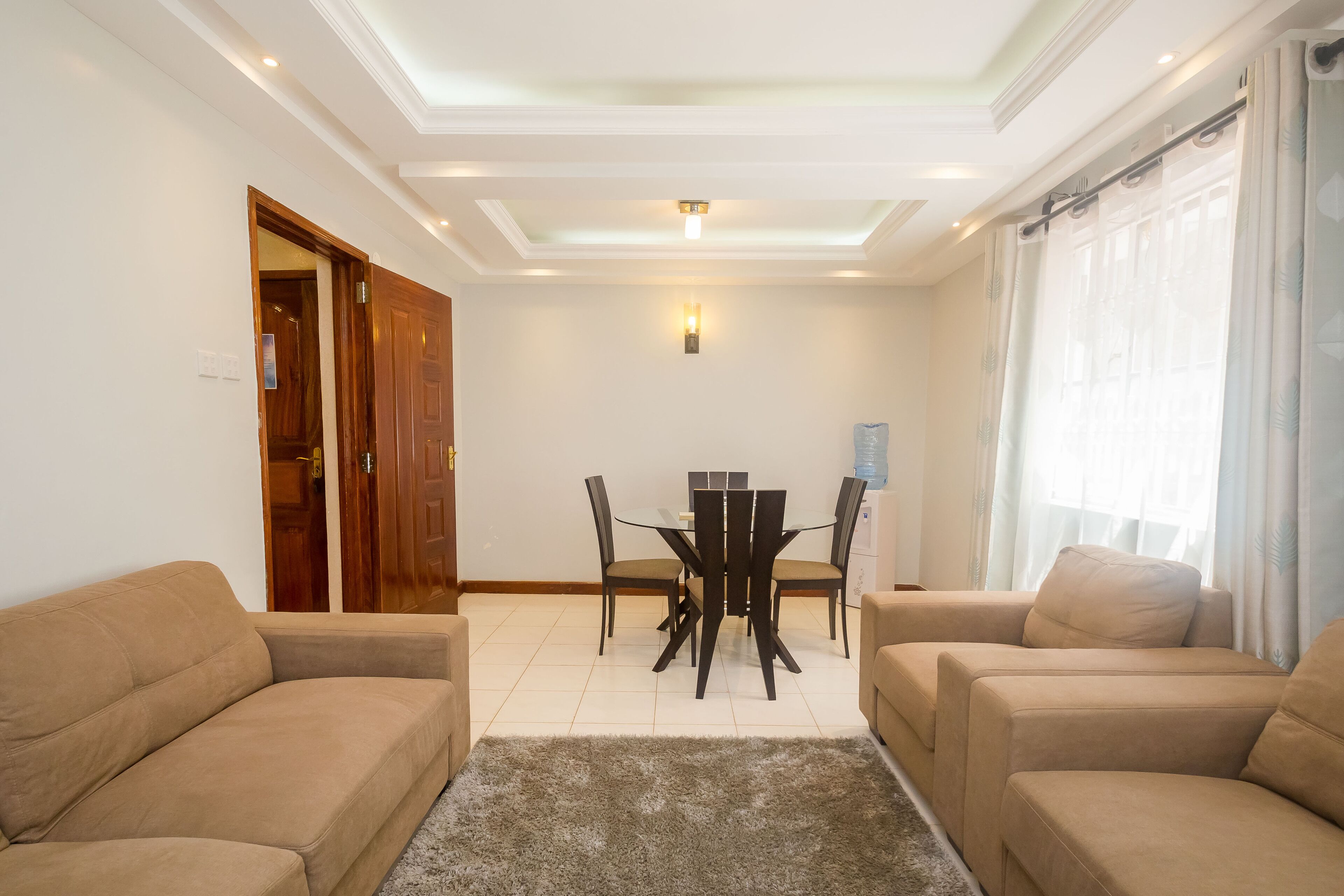 A living room with beige sofas, a glass-top dining table, and a ceiling light fixture.