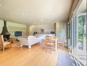 A modern living room with a fireplace, a sofa, a dining table with chairs, a kitchen area, and large windows.