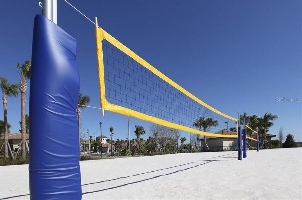 Una cancha de voleibol de playa con red, postes azules y amarillos, y palmeras al fondo.