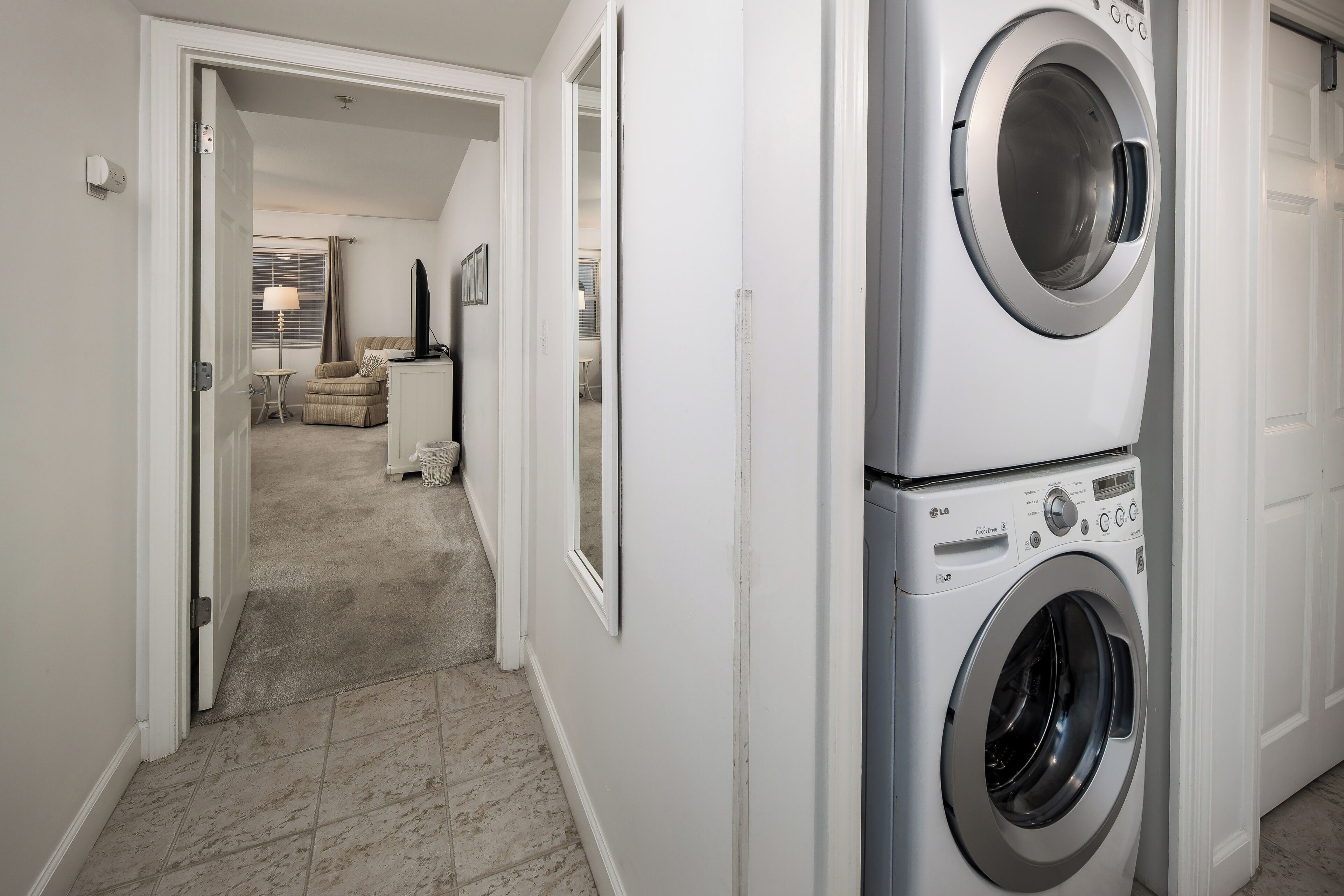 A laundry room with stacked washers and dryers, a hallway leading to a living area with a couch and lamp, and a door to another room.