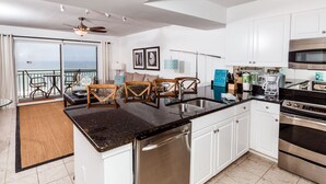 A modern kitchen with white cabinets, a stainless steel dishwasher, and an oven. There is a dining area with a table and chairs, and a balcony with a view of the ocean. - Pelican Isle 507 - Breezy Bliss (Fort Walton Beach)