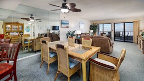 A spacious living area with a dining table, wicker furniture, and a balcony with a view. - Surf Dweller 709 - Tidal Treasures (Fort Walton Beach)