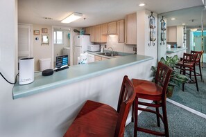 A compact kitchen with wooden cabinets, a microwave, and a sink. There are two wooden chairs and a small table. - Surf Dweller 709 - Tidal Treasures (Fort Walton Beach)