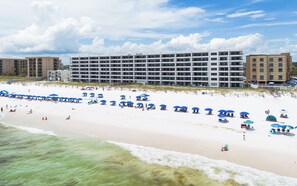 A beachfront hotel with multiple floors, a sandy beach with blue umbrellas, and a clear sky. - Surf Dweller 409 - Vista Vibe (Fort Walton Beach)