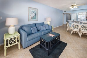 A living room with a blue sofa, two side tables with lamps, a coffee table, and a dining area with wicker chairs and a white table. - Emerald Towers West 2005 - Emerald Shore (Fort Walton Beach)