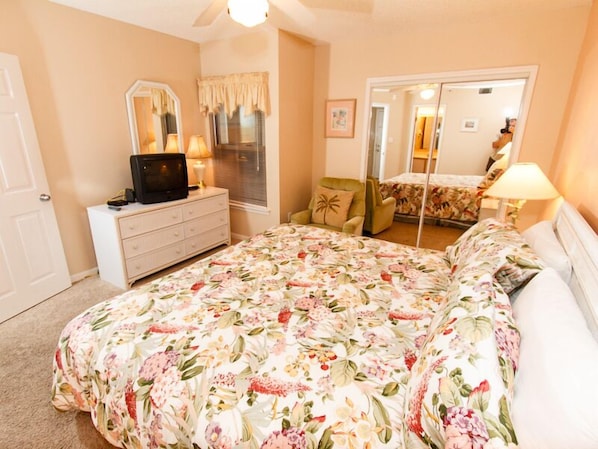 A bedroom with a floral bedspread, a dresser with a television, a chair, and a mirror reflecting the room. - Emerald Towers West 2005 - Emerald Shore (Fort Walton Beach)