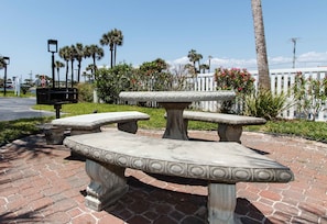A stone picnic table with a bench, surrounded by palm trees and flowering plants. - Emerald Twrs West 3003 by Brooks and Shorey Resorts (Fort Walton Beach)