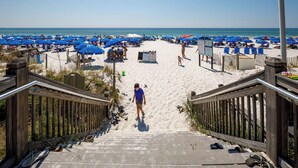 A beach with a wooden staircase leading down to the sand, surrounded by blue umbrellas and beach chairs. - Waterscape B310 - Resort Shores (Fort Walton Beach)