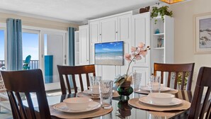 A dining area with a glass table, wooden chairs, and a view of the ocean through large windows. - Gulfside 302 - Ocean Serenity (Fort Walton Beach)