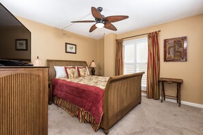 A bedroom with a wooden bed, a ceiling fan, a small table, and a window with curtains. - Gulfside 501 - Sandy Adventures (Fort Walton Beach)