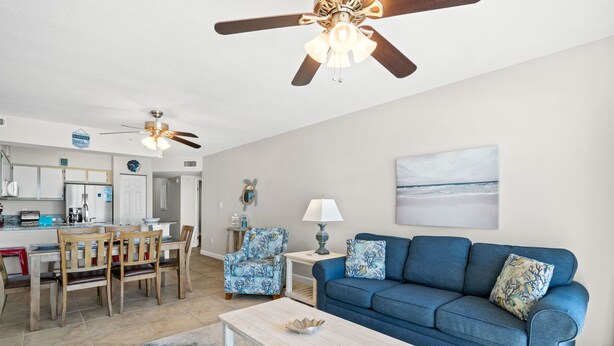 Ein modernes Wohnzimmer mit einem blauen Sofa, einem Holzespeisesaal und Stühlen, einer Küchenzeile mit Kühlschrank und Herd sowie einem Deckenventilator.