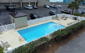 A swimming pool with a white fence, lounge chairs, and a parking lot with various vehicles. - Emerald Towers West 1003 - Sandy Toes (Fort Walton Beach)